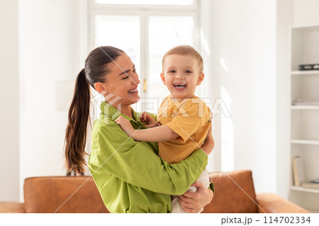 A woman is standing, holding a child in her arms. Little son appears to be resting against her chest, while the mother gazes down at them lovingly 114702334