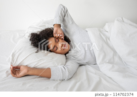 Hispanic woman is laying on a bed with white sheets. The room is simple and tidy, with natural light streaming in, waking up. The lady appears relaxed and comfortable, her eyes closed as she rests. Hispanic woman is laying on a bed with white sheets. The room is simple and tidy, with natural light streaming in, waking up. The lady appears relaxed and comfortable, her eyes closed as she rests. 114702374