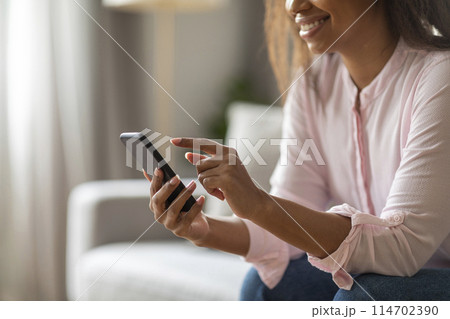 Enjoying a quiet day indoors, black woman sits on her couch using her mobile phone, surrounded by a tastefully decorated living room in soft colors, cropped 114702390