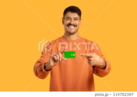 A man wearing an orange shirt is seen holding a green card in his hand on yellow studio background, contactless payment A man wearing an orange shirt is seen holding a green card in his hand on yellow studio background, contactless payment 114702397