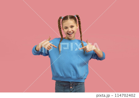 Cheerful young girl with pigtails in a blue sweater confidently pointing at herself against a pink background 114702402