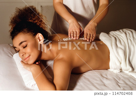 An African American woman is depicted receiving a back scrub treatment, emphasizing the themes of luxury spa care and relaxation for people 114702408