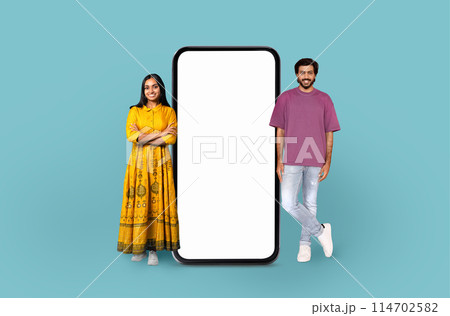 Indian woman in traditional dress and a man in casual wear stand beside a large blank smartphone screen, implying technology use 114702582