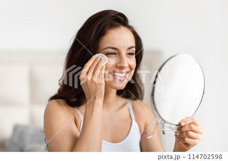 Cheerful millennial lady using a cotton pad for skin care as she looks in the mirror in a bathroom 114702598