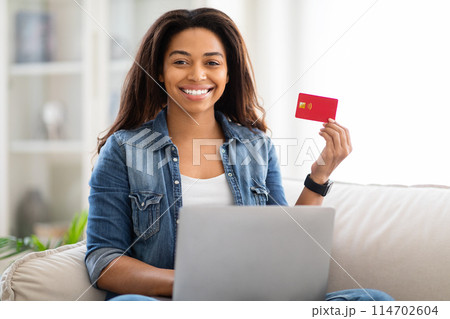 African American woman is seated on a couch, holding a credit card and a laptop in her hands, engaging in online shopping or financial transactions African American woman is seated on a couch, holding a credit card and a laptop in her hands, engaging in online shopping or financial transactions 114702604