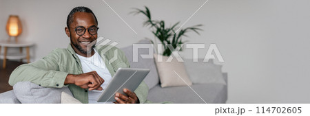 Smiling mature african american man in glasses and casual watching video on tablet, chatting in social networks in living room interior. New app, work remote at home, blog, panorama with copy space Smiling mature african american man in glasses and casual watching video on tablet, chatting in social networks in living room interior. New app, work remote at home, blog, panorama with copy space 114702605