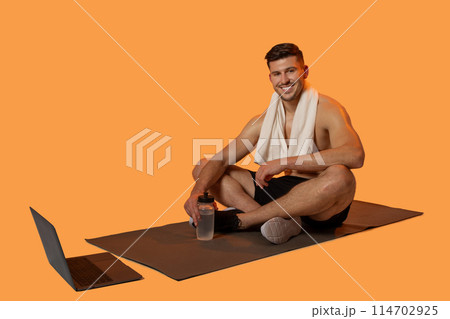A cheerful man sits on a yoga mat with a white towel draped over his shoulders, holding a water bottle. With a laptop beside him, it appears he following an online workout or yoga session 114702925