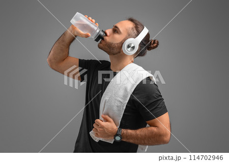 A man is seen wearing headphones as he takes a sip from a water bottle. He appears to be enjoying a refreshment while listening to music through his headphones. 114702946