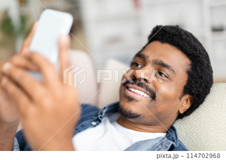 An african american guy enjoys browsing on his smartphone, exuding happiness and relaxation in a comfortable home setting 114702968