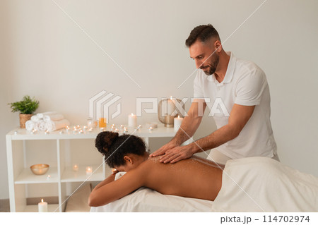 A professional masseur provides a relaxing back massage to black woman in a serene spa setting with candles and towels A professional masseur provides a relaxing back massage to black woman in a serene spa setting with candles and towels 114702974