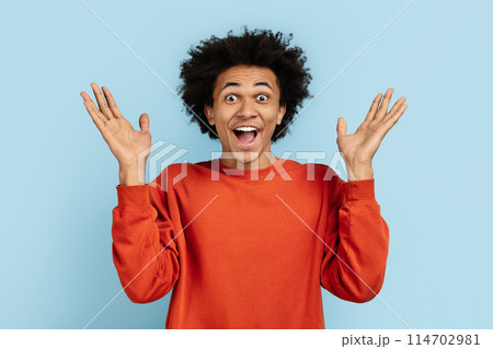 African american man in an orange sweater looking shocked with raised hands, isolated on a blue background, portraying surprise and excitement 114702981