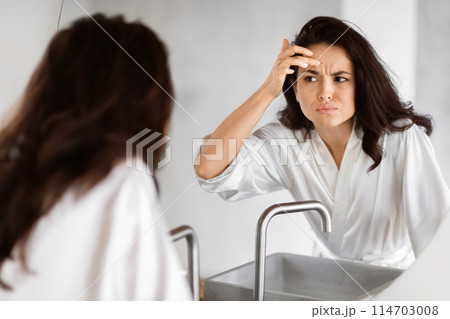A woman in a white robe appears concerned as she examines her forehead for wrinkles in the bathroom mirror, embodying beauty anxieties A woman in a white robe appears concerned as she examines her forehead for wrinkles in the bathroom mirror, embodying beauty anxieties 114703008