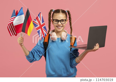 A tech-savvy teen girl showcasing a laptop while holding various national flags, isolated on a pink backdrop 114703019