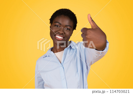 A smiling young african american woman with short hair gives a thumbs up sign against a bright yellow background, expressing positivity A smiling young african american woman with short hair gives a thumbs up sign against a bright yellow background, expressing positivity 114703021