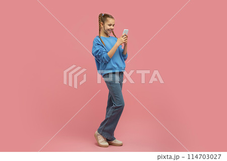 A cheerful young girl with braided hair is standing, happily looking at her smartphone against a pink background 114703027