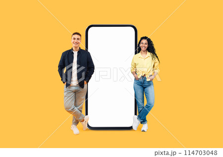 Smiling young man and woman posing beside enormous smartphone display with mockup copy space on a yellow background 114703048