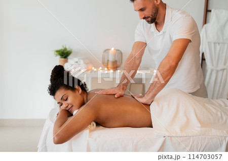 A skilled masseur administers a relaxing back massage to an african american lady at a modern spa, emphasizing human touch and professional care 114703057