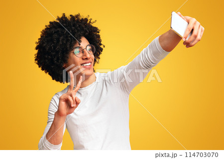 Young black guy making selfie with peace gesture on cellphone, orange studio background 114703070
