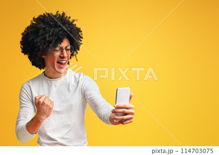 Success. Emotional african guy reading message on cellphone and gesturing with fist, orange studio background Success. Emotional african guy reading message on cellphone and gesturing with fist, orange studio background 114703075