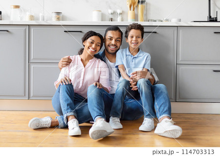 Black parents and child laughing together while seated on the kitchen floor, displaying a relaxed family atmosphere 114703313
