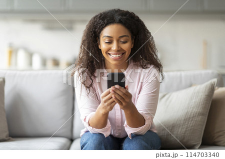 Smiling african american lady enjoying time on her smartphone at home, showcasing millennials' digital lifestyle 114703340