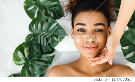 This vibrant image shows a black woman smiling at a spa, surrounded by green leaves, with a hand gently framing her face, evoking happiness 114703356