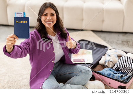 A middle eastern woman sitting on the floor with an open suitcase and clothes gives a thumbs up, holding her passport, ready to travel 114703363