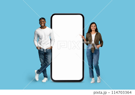 Smiling Multiracial couple pointing towards a huge mobile phone illustration on blue studio background 114703364