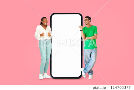 African American man giving thumbs up and woman smiling beside an empty screen on a pink backdrop, ideal for advertisements 114703377