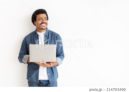 A cheerful man in casual denim attire holding a laptop computer and looking to the side with a hopeful expression, copy space 114703400