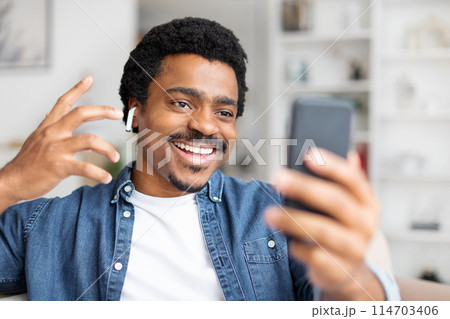 A cheerful young black man wearing earbuds smiles as he looks at his smartphone, possibly engaging in a video call or taking a selfie 114703406
