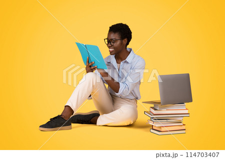 An african american zoomer woman absorbed in reading against an isolated yellow background with a laptop and a stack of books, reflecting a love for learning An african american zoomer woman absorbed in reading against an isolated yellow background with a laptop and a stack of books, reflecting a love for learning 114703407
