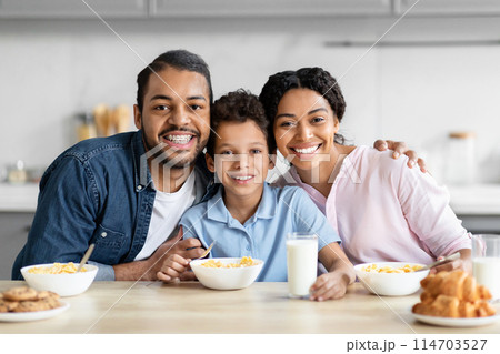 A joyful african american family with a father, mother, and son are seated at the kitchen table, enjoying breakfast and spending quality time together 114703527