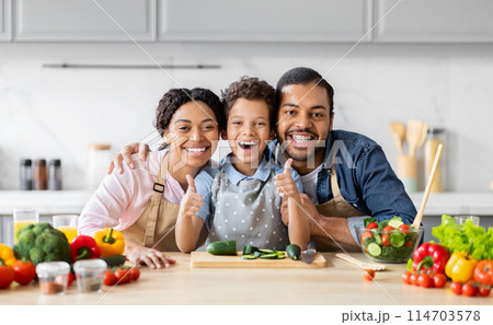 Energetic African American family posing with thumbs up in the kitchen, portraying a fun family cooking moment with healthy food Energetic African American family posing with thumbs up in the kitchen, portraying a fun family cooking moment with healthy food 114703578