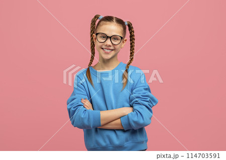 A cheerful young girl with braided pigtails and eyeglasses stands confidently with arms crossed on a pink background A cheerful young girl with braided pigtails and eyeglasses stands confidently with arms crossed on a pink background 114703591
