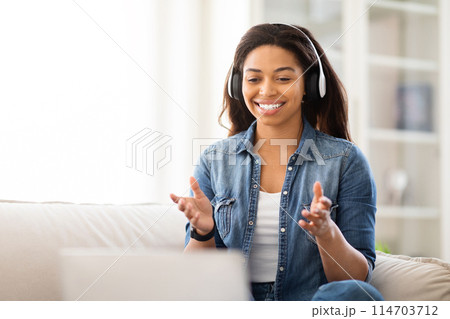 African American woman sitting on a couch with headphones on, listening to music or a podcast. She appears relaxed and focused on the sound coming from her headphones. 114703712