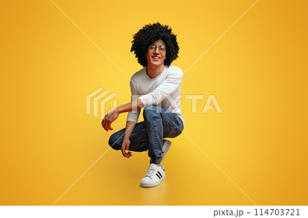 My style. Studio portrait of young black man hunkering down and smiling, orange background 114703721