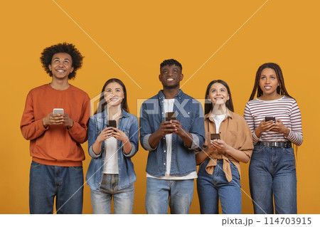 A diverse international team of young friends engaged with their mobile phones, representing a multiethnic, multiracial generation isolated on an orange backdrop A diverse international team of young friends engaged with their mobile phones, representing a multiethnic, multiracial generation isolated on an orange backdrop 114703915