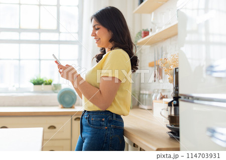 Illustrates a cozy home scene, perhaps involving a middle eastern woman, in a sunlit kitchen engaging with her smartphone Illustrates a cozy home scene, perhaps involving a middle eastern woman, in a sunlit kitchen engaging with her smartphone 114703931