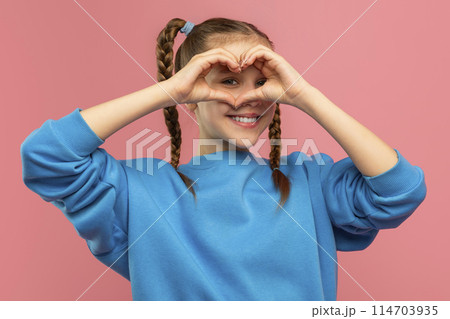 Cute teen girl showing heart gesture over her face and smiling, posing isolated on pink studio background 114703935