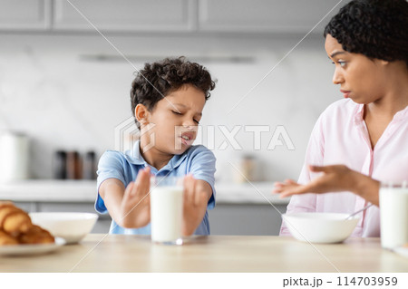 The image illustrates African American boy refusing a glass of milk with a grimace, showcasing a child's aversion or dietary choices in a family kitchen 114703959