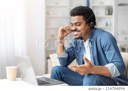 A cheerful young black man wearing headphones is engaged in a video call or remote work on his laptop in a bright room A cheerful young black man wearing headphones is engaged in a video call or remote work on his laptop in a bright room 114704054