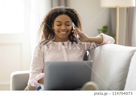 The African American woman is smiling while she talks on the phone and looks at her laptop, showing a home multitasking scenario 114704073