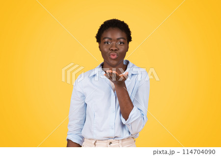 Playful and charismatic black woman sends a flying kiss towards the camera, highlighting her liveliness and Gen Z lifestyle, isolated yellow setting Playful and charismatic black woman sends a flying kiss towards the camera, highlighting her liveliness and Gen Z lifestyle, isolated yellow setting 114704090