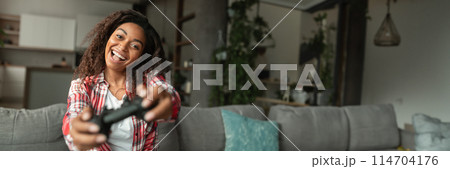 Glad excited young african american woman with open mouth playing online game with joystick, sitting on sofa in living room interior, close up. Fun at home alone, entertainment, copy space 114704176