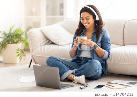 African American woman is sitting cross-legged on the floor in a room. She is focused on her black laptop screen, drinking coffee while watching video African American woman is sitting cross-legged on the floor in a room. She is focused on her black laptop screen, drinking coffee while watching video 114704212