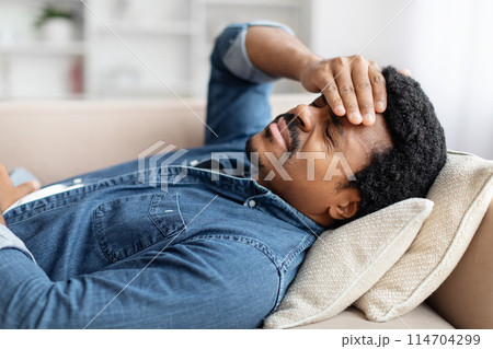 Captured in a serene home environment, this image portrays a young african american guy lying on couch at home, touching head, suffering from migraine Captured in a serene home environment, this image portrays a young african american guy lying on couch at home, touching head, suffering from migraine 114704299