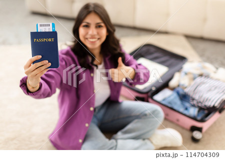 A smiling young middle eastern woman is sitting on the floor with an open suitcase, holding a passport in one hand and giving a thumbs up with the other 114704309