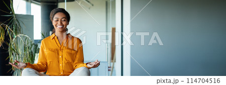 Office zen. Calm black businesswoman meditating with closed eyes on office desk, sitting at workplace in lotus position. Positive woman feeling peaceful and balanced, panorama with copy space 114704516
