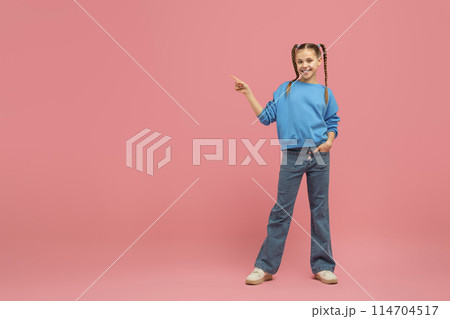 A cheerful young girl with braided pigtails, wearing a blue sweater and jeans, pointing to her side against a vibrant pink background, copy space A cheerful young girl with braided pigtails, wearing a blue sweater and jeans, pointing to her side against a vibrant pink background, copy space 114704517